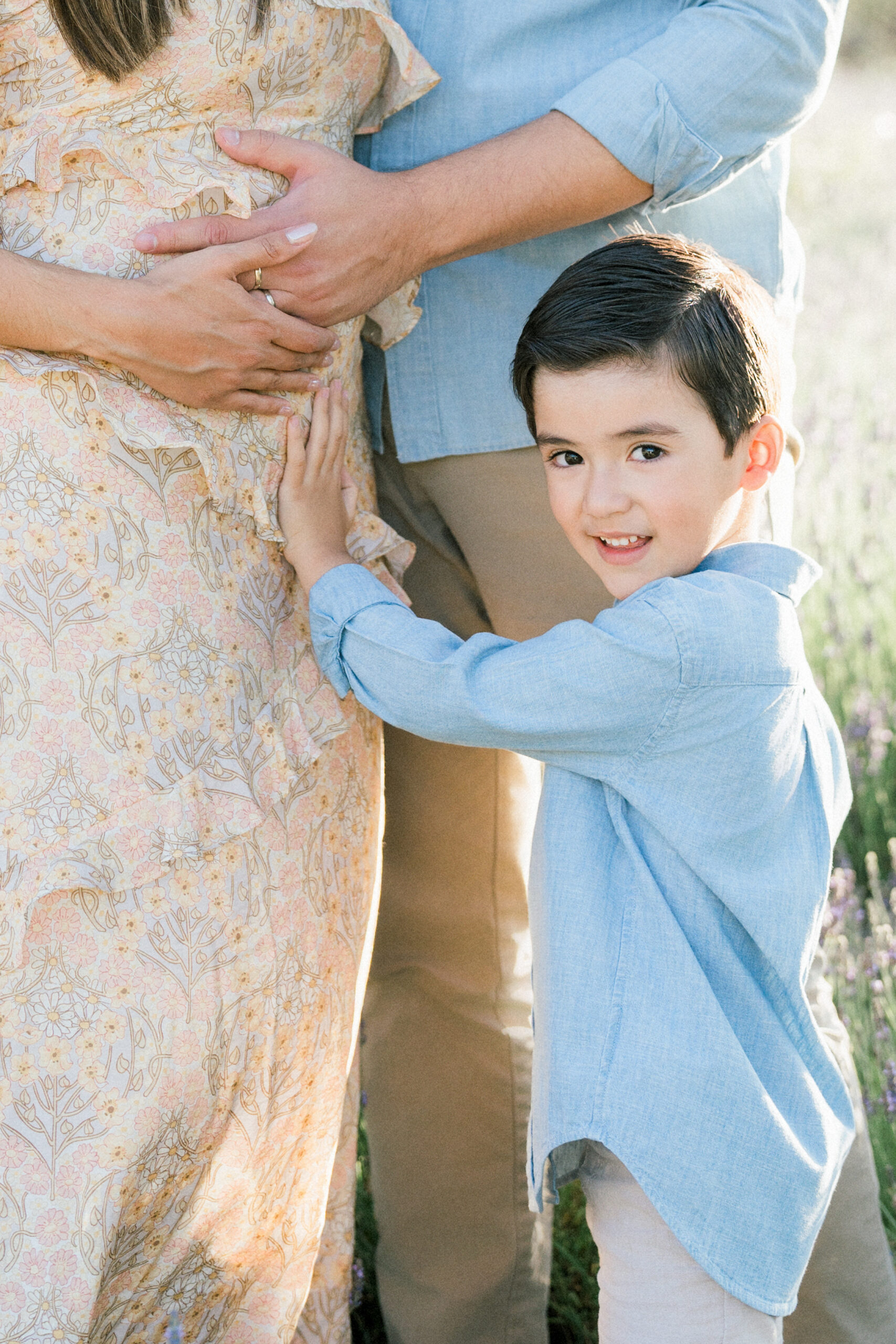 Big Bro feeling pregnant belly while smiling at the camera with lavender fields in the background. Taken by a maternity photographer.